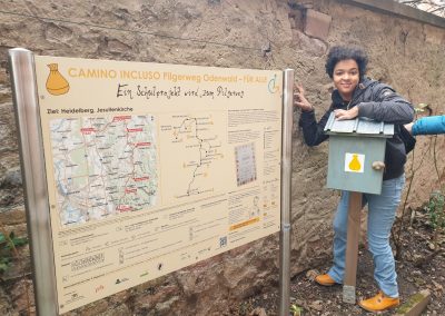 Marlene kontrolliert den Pilger · kasten. Marlene steht vor der letzten Info · tafel am Ziel vom Camino Incluso.